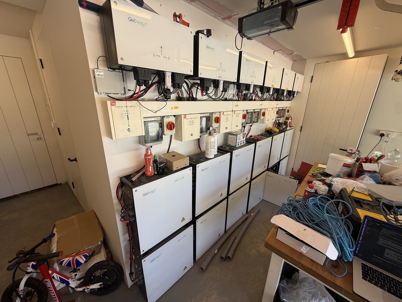 A garage plant room with a long row of GivEnergy AC3 inverters mounted across the wall, a bank of labelled isolators and essential-power-supply distribution boards beneath them, and a stack of white GivEnergy battery modules below — one of the largest residential GivEnergy installations in the country.
