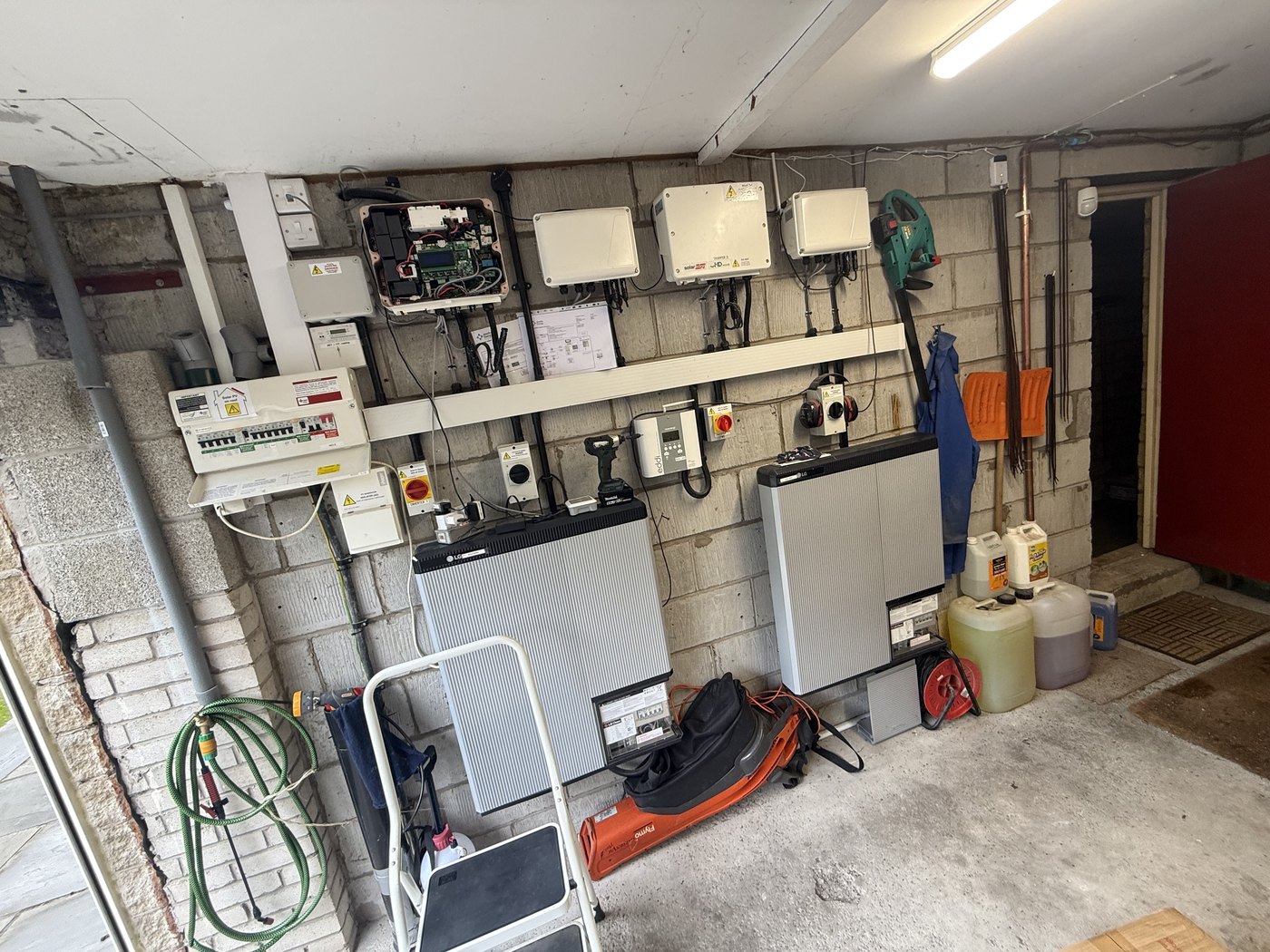 A garage plant room with a wall of SolarEdge inverters and two LG Chem battery units mounted on a block wall, alongside the consumer unit, isolators and meter — a premium solar-plus-storage system installed about ten years ago.